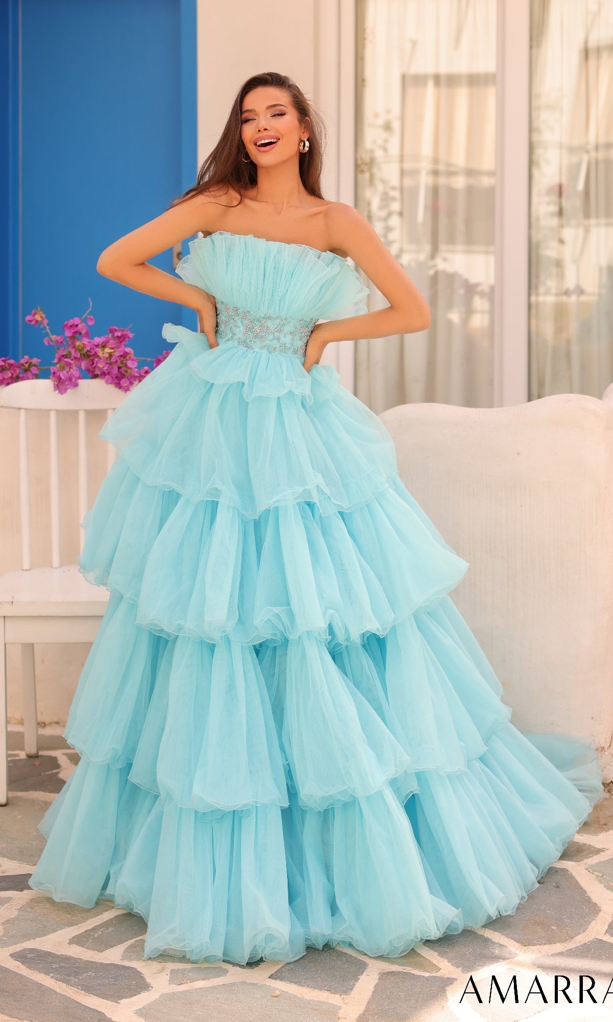 A woman poses outdoors, smiling with one hand on her hip, wearing the Amarra Long Formal Dress 88083 featuring a strapless beaded waist and layered light blue tulle skirt.